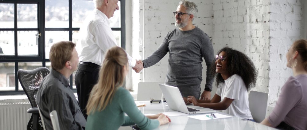 An employer shakes an employees hand at a conference table with other employees.